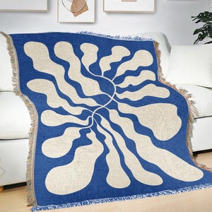 May include: A blue throw blanket with a cream abstract pattern. The blanket, with fringed edges, is draped over a white sofa. The design features a central sun-like shape with flowing, organic lines radiating outwards, creating a modern aesthetic.