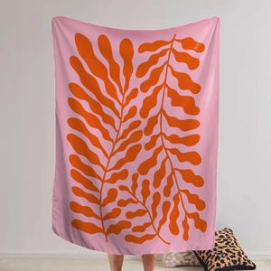 May include: A pink blanket with a large orange leaf design. The leaves are stylized and symmetrical, creating a bold, graphic pattern. The blanket is soft and appears to be made of a comfortable material. The overall aesthetic is modern and artistic.