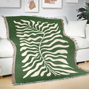 May include: A green throw blanket with a white leaf design draped over a white sofa. The blanket has a fringed edge and a repeating pattern of a stylized leaf. The background includes framed art and a houseplant.