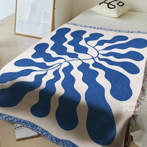 May include: A woven throw blanket with a cream background and a large, stylized blue floral design. The blanket has a fringed edge and is draped over a bed. The design is reminiscent of a botanical illustration.