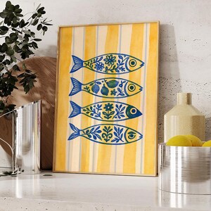 May include: A framed art print featuring four stylized fish with blue and yellow floral patterns. The background has vertical yellow stripes. The artwork is displayed on a white countertop with other decorative items.
