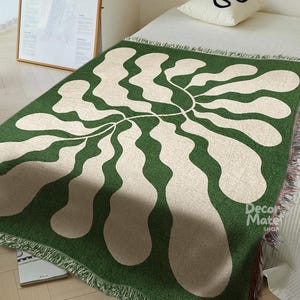 May include: Green and cream woven throw blanket with an abstract floral pattern. The blanket has a fringed edge and is draped over a bed. The design features a central stem with flowing, organic shapes. The blanket is made of a soft material.