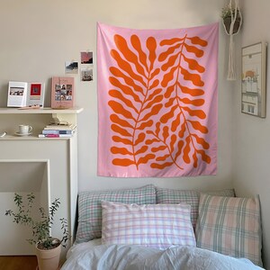 May include: A pink tapestry with a large orange leaf design hangs on a white wall. The tapestry is the focal point of a bedroom scene, with a bed covered in pillows and a blanket. A small plant in a pot sits on the floor.