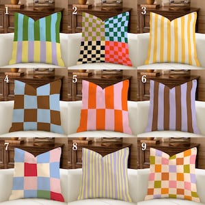 Peut inclure: Neuf coussins décoratifs avec divers motifs géométriques et couleurs. Les coussins présentent des rayures, des carreaux et des motifs blocs de couleurs dans des tons de bleu, vert, jaune, orange, rose, marron et violet. Les coussins sont disposés sur un canapé blanc.