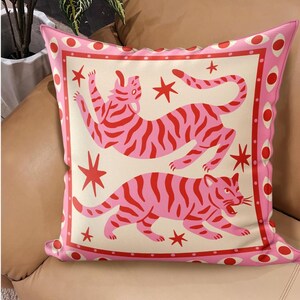 May include: Pink decorative pillow with a cream-colored center featuring two stylized pink tigers with red stripes and red stars. The pillow has a pink border with red and white circular accents, creating a playful and vibrant design.