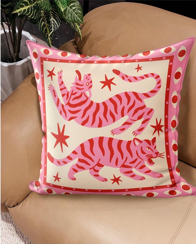 May include: Pink decorative pillow with a cream-colored center featuring two stylized pink tigers with red stripes and red stars. The pillow has a pink border with red and white circular accents, creating a playful and vibrant design.