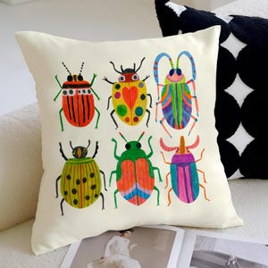 May include: Cream-colored square throw pillow featuring a colorful illustration of six whimsical beetles. The beetles are in various colors, including red, yellow, green, and blue, with detailed patterns. The pillow is placed on a white sofa.