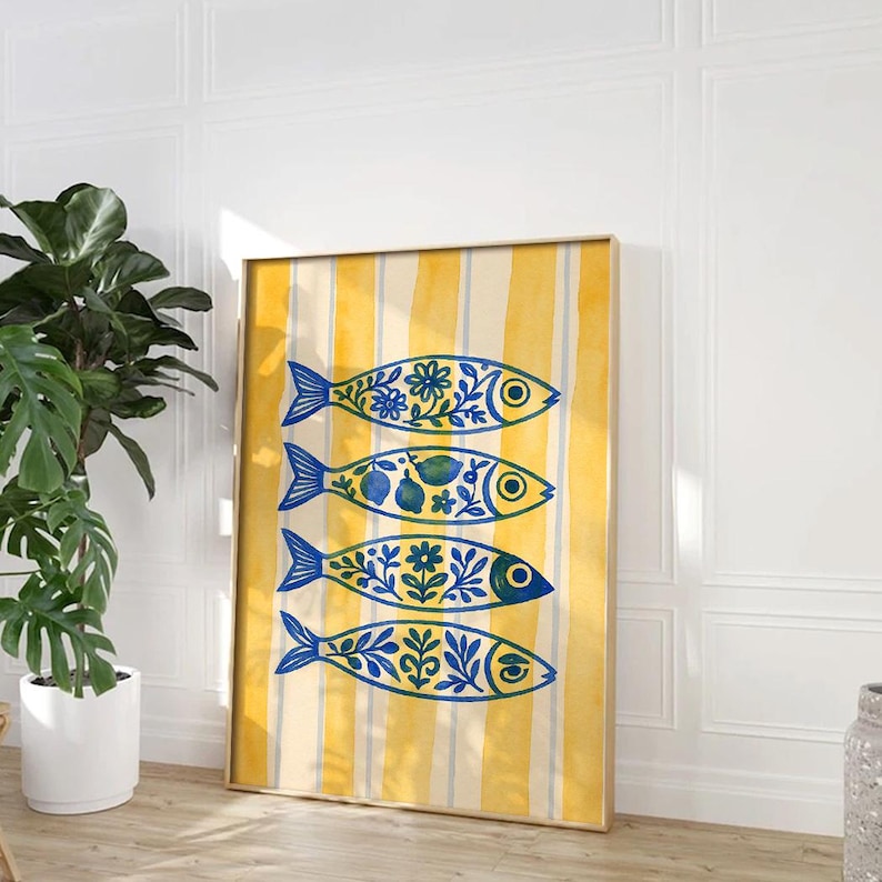 May include: A framed art print featuring four stylized fish in blue and yellow. Each fish is decorated with floral and fruit patterns. The background has vertical yellow and white stripes. The artwork is displayed in a room with a plant.