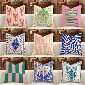 Puede incluir: Nueve cojines decorativos con varios diseños se muestran en un sofá blanco. Los cojines presentan patrones como langostas, conchas marinas, peces, corazones, texto y formas geométricas. Los colores incluyen rosa, verde, azul y crema.