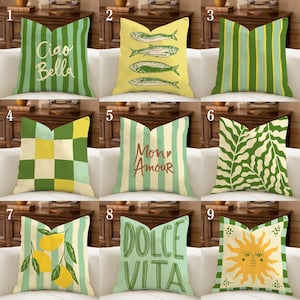 May include: A collection of decorative throw pillows with various designs. Pillows include vertical stripes, fish illustrations, checkered patterns, botanical prints, and text such as "Ciao Bella", "Mon Amour", and "Dolce Vita". Colors include green, yellow, and cream.