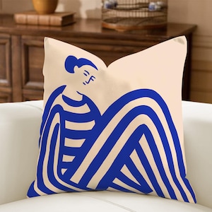 May include: A square cushion featuring a cream background and a blue abstract illustration of a person. The person has a simple face and is wearing a striped top and has striped legs. The cushion is made of a soft fabric.