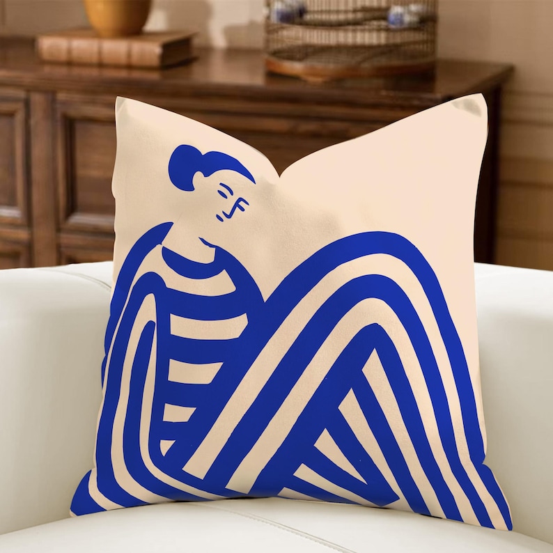 May include: A square cushion featuring a cream background and a blue abstract illustration of a person. The person has a simple face and is wearing a striped top and has striped legs. The cushion is made of a soft fabric.