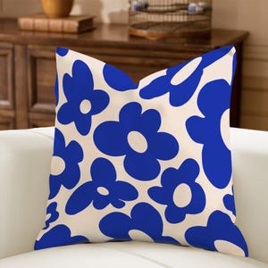 May include: A square throw pillow with a cream-colored background and a repeating pattern of large, stylized blue flowers. The pillow is placed on a white sofa, suggesting a home decor item.