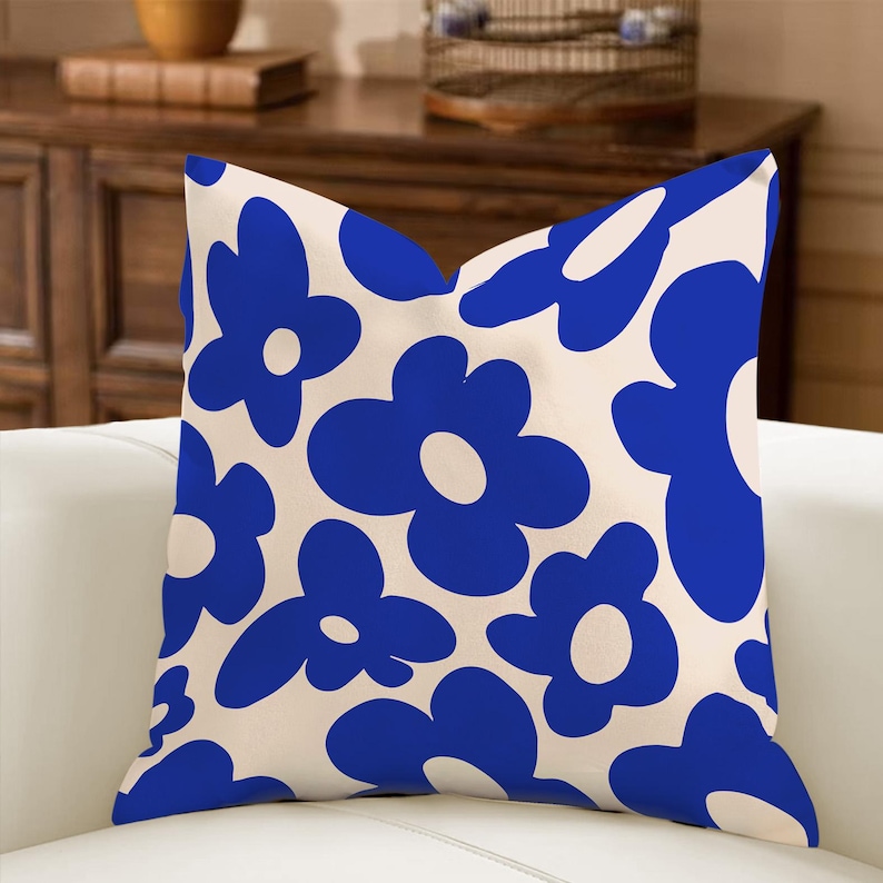 May include: A square throw pillow with a cream-colored background and a repeating pattern of large, stylized blue flowers. The pillow is placed on a white sofa, suggesting a home decor item.