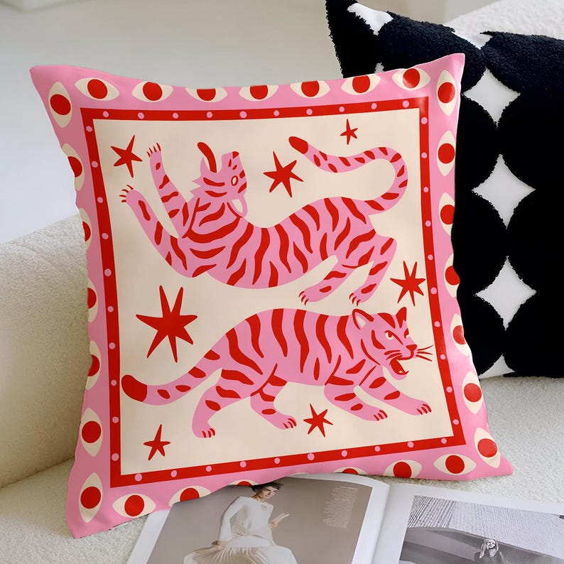 May include: A decorative square pillow with a pink border featuring red eye-like shapes. The pillow's center has a cream background with two pink and red tiger illustrations and red star shapes.