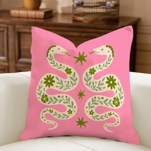 May include: A pink decorative pillow featuring two stylized snakes facing each other. The snakes are cream-colored with green floral and leaf patterns. Green star and flower accents are scattered across the pink background.