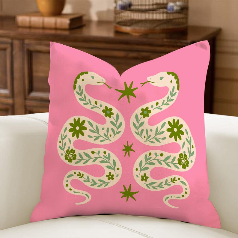 May include: A pink decorative pillow featuring two stylized snakes facing each other. The snakes are cream-colored with green floral and leaf patterns. Green star and flower accents are scattered across the pink background.