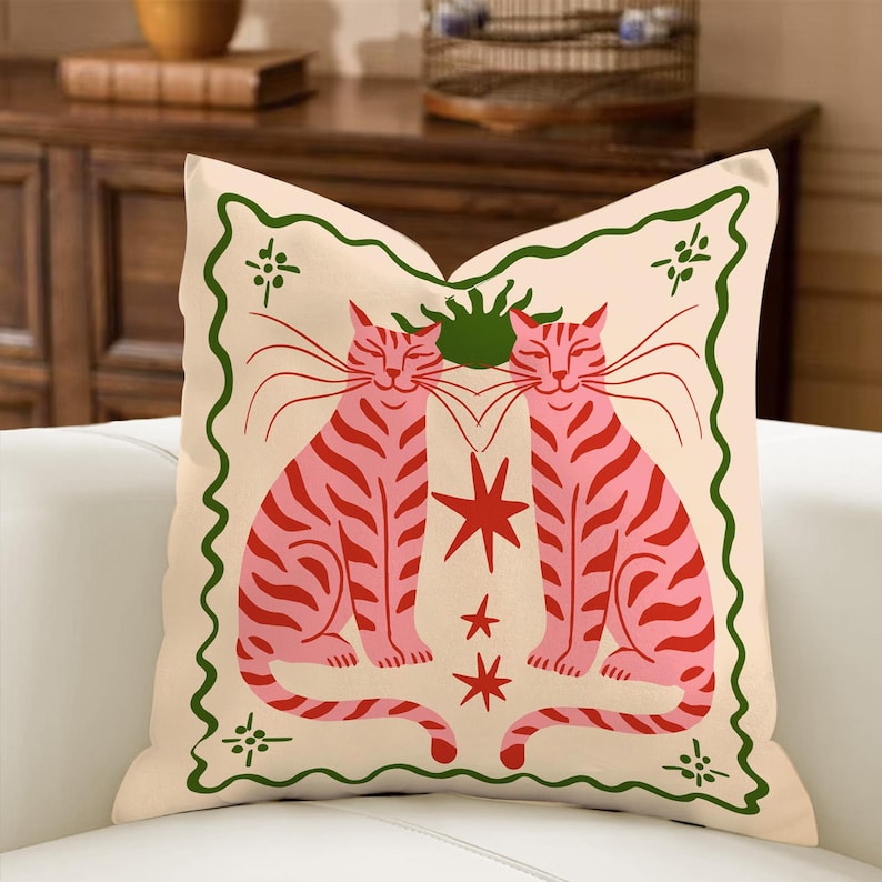 May include: Cream-colored decorative pillow featuring two pink and red striped cats facing each other. The design includes a green border with floral accents and red star shapes. The pillow is on a white couch.