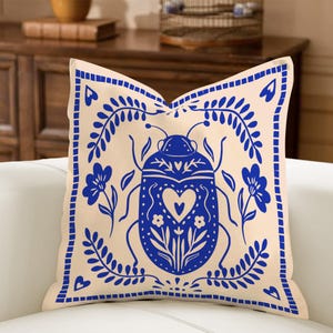 May include: Beige square throw pillow with a blue insect and floral design. The insect has a heart shape in the center. The pillow has a blue border with a floral pattern. Decorative pillow for a sofa or bed.