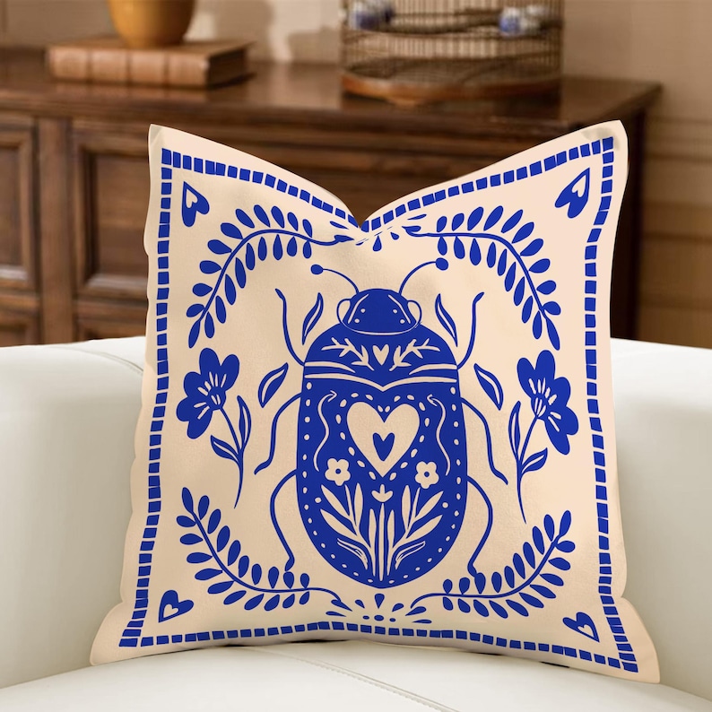 May include: Beige square throw pillow with a blue insect and floral design. The insect has a heart shape in the center. The pillow has a blue border with a floral pattern. Decorative pillow for a sofa or bed.