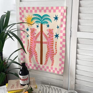 Puede incluir: Un tapiz decorativo con un dise&ntilde;o caprichoso. Presenta dos tigres rosas flanqueando una palmera. El fondo tiene un patr&oacute;n de cuadros rosas y blancos, con un sol amarillo y estrellas azules.