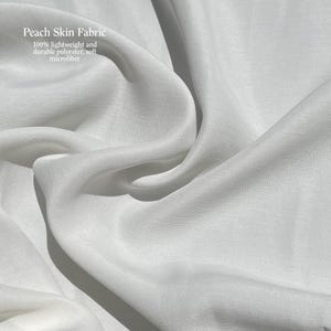 May include: Close-up of white peach skin fabric with soft, flowing folds. The fabric is 100% lightweight and durable polyester microfiber. The text "Peach Skin Fabric" is visible in the upper left corner.
