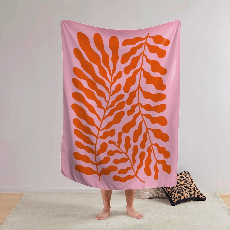 May include: A pink blanket with a large orange leaf design. The leaves are stylized and symmetrical, creating a bold, graphic pattern. The blanket is soft and appears to be made of a comfortable material. The overall aesthetic is modern and artistic.