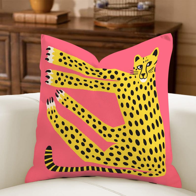 May include: A square cushion with a pink background and a yellow cheetah design. The cheetah has black spots and elongated limbs. The cushion is on a white surface. The design is modern and playful.