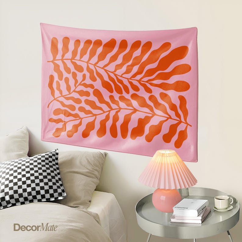 May include: A pink wall tapestry with a large orange leaf design. The tapestry is hanging above a bed with pillows and a side table with a lamp and books. The tapestry is a decorative home decor item.