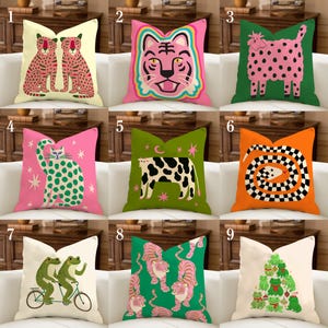 Puede incluir: Una colección de cojines decorativos con varios diseños de animales y naturaleza. Los cojines presentan ilustraciones de tigres, gatos, vacas, ranas y serpientes en colores vibrantes como rosa, verde, naranja y amarillo. Los cojines están colocados en un sofá blanco.