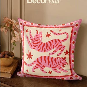May include: Pink decorative pillow featuring two stylized tigers with red stripes and stars on a cream background. The pillow has a pink border with red circles and the text "DecorMate" at the top.