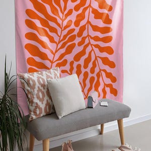 May include: A pink wall tapestry with a large orange leaf design hangs above a gray bench with pillows and a small purse. A potted plant and a rug with a geometric pattern are in the foreground. The scene is a modern interior.
