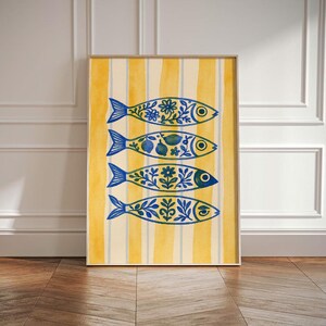 May include: A framed art print featuring four blue fish with intricate floral and fruit designs. The fish are set against a yellow and white striped background. The artwork is in a wooden frame and leans against a white wall.