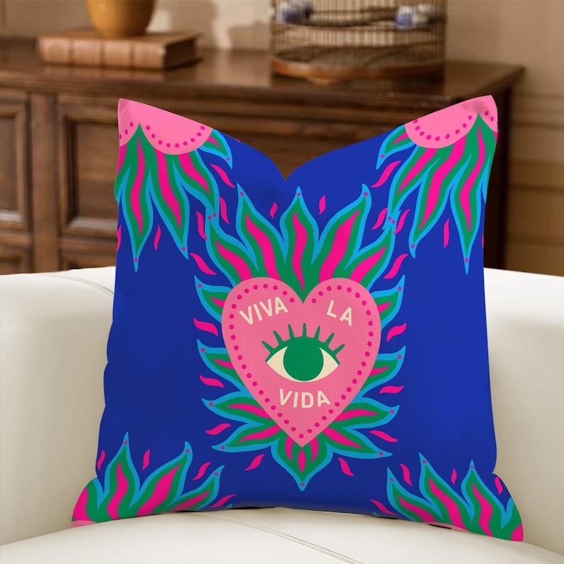 May include: A square blue decorative pillow with a pink heart-shaped design in the center. The heart contains an eye and the words "VIVA LA VIDA". The design is surrounded by pink, green, and blue flame-like shapes.