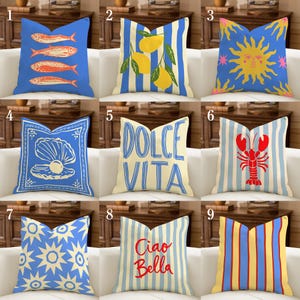 Può includere: Una collezione di cuscini decorativi con vari disegni. I cuscini presentano illustrazioni di pesci, limoni, un sole, una conchiglia, le parole "Dolce Vita", un'aragosta, scoppi di stelle, le parole "Ciao Bella" e strisce verticali. I cuscini sono su un divano bianco.