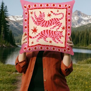 May include: Pink decorative pillow with a cream-colored center featuring two pink striped tigers and red star accents. The pillow has a pink border with red eye and dot designs. The text "DecorMate" is at the top.