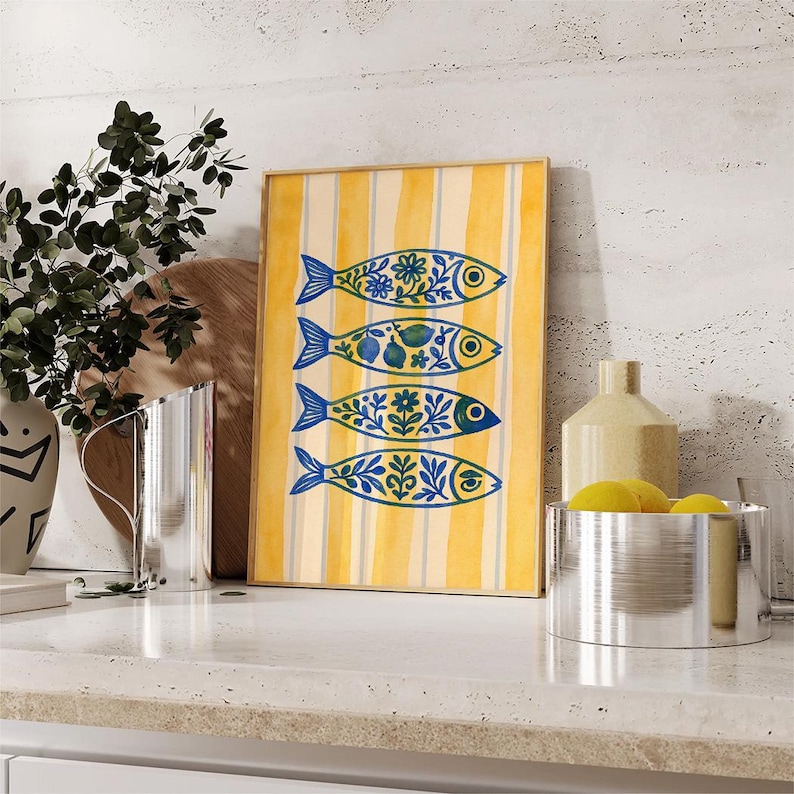 May include: A framed art print featuring four stylized fish with blue and yellow floral patterns. The background has vertical yellow stripes. The artwork is displayed on a white countertop with other decorative items.