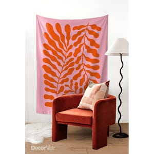 May include: A pink wall tapestry with a large orange leaf design hangs behind a rust-colored armchair. A patterned pillow rests on the chair. A black, wavy floor lamp stands to the right. A white rug is on the floor.