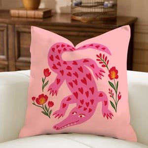 May include: Pink cushion featuring a pink alligator covered in red hearts. The alligator is surrounded by red flowers with green stems and leaves, set against a solid pink background.