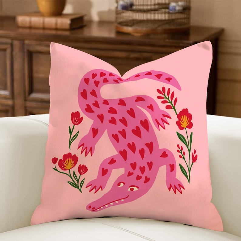 May include: Pink cushion featuring a pink alligator covered in red hearts. The alligator is surrounded by red flowers with green stems and leaves, set against a solid pink background.