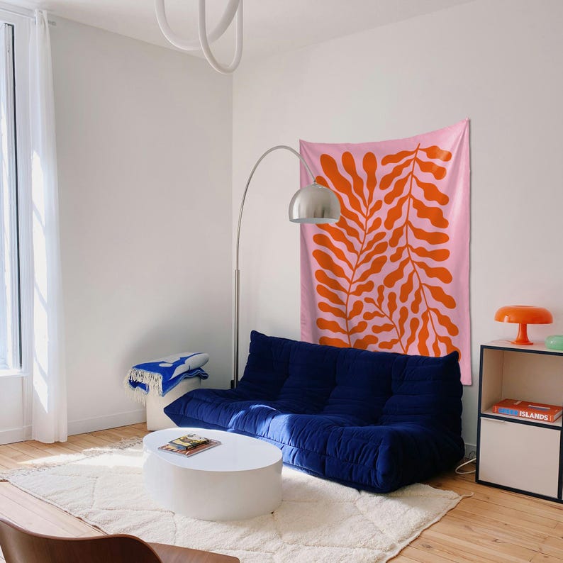 May include: A modern living room with a navy blue modular sofa, a white coffee table, and a pink wall hanging with orange leaf-like patterns. A silver arc lamp and a small orange lamp add to the decor. The room has a white rug and hardwood floors.
