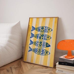 May include: A framed art print featuring four stylized blue fish with floral and fruit patterns against a yellow and white striped background. The print is leaning against a white wall. The DecorMate logo is visible in the top left corner.