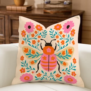 May include: Beige square throw pillow with a colorful insect and floral design. The insect is pink and orange with black antennae and legs. The flowers are pink and orange, and the leaves are teal.
