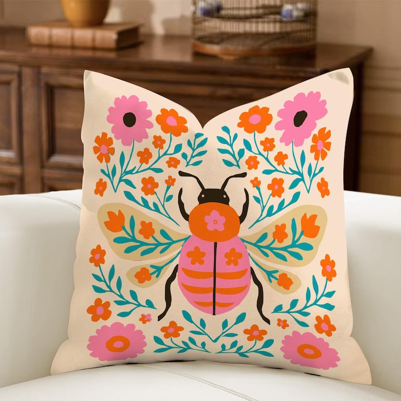 May include: Beige square throw pillow with a colorful insect and floral design. The insect is pink and orange with black antennae and legs. The flowers are pink and orange, and the leaves are teal.