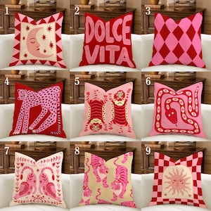 Pode incluir: Uma coleção de nove almofadas quadradas com vários padrões em vermelho e rosa, incluindo uma lua crescente, o texto Dolce Vita, formas de diamante, um leopardo, tigres, uma cobra, leões e um sol, dispostos num sofá branco.