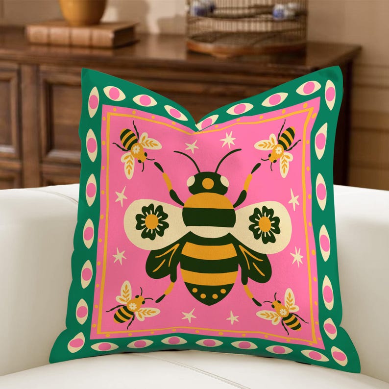 May include: A decorative square pillow with a pink center featuring a large bee illustration with yellow and black stripes, surrounded by smaller bees and stars. The pillow has a green border with pink oval accents, creating a vibrant and whimsical design.