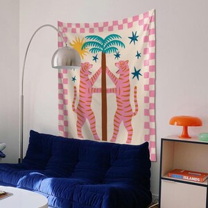 Puede incluir: Un tapiz de pared decorativo con dos tigres rosas abrazando una palmera, con un sol y estrellas sobre un fondo a cuadros rosa y blanco. El tapiz cuelga sobre un sof&aacute; azul en un ambiente de sala de estar.