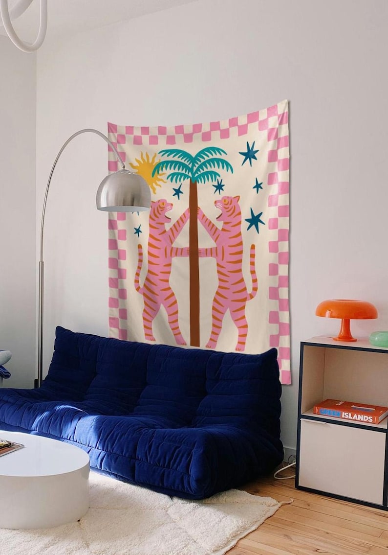 Puede incluir: Un tapiz de pared decorativo con dos tigres rosas abrazando una palmera, con un sol y estrellas sobre un fondo a cuadros rosa y blanco. El tapiz cuelga sobre un sof&aacute; azul en un ambiente de sala de estar.
