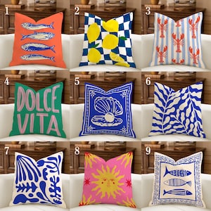 Puede incluir: Una colección de cojines decorativos con varios diseños. Los cojines presentan imágenes de peces, limones, langostas, las palabras "Dolce Vita", una concha, formas azules abstractas, un sol y más peces. Los cojines son de varios colores, incluyendo naranja, azul, verde, rosa y blanco.