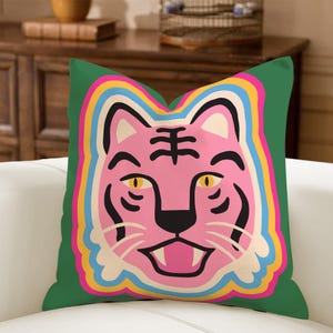 May include: A square cushion with a green background. It features a pink tiger face with black stripes and yellow eyes. The tiger face is outlined with a rainbow of colours, including pink, yellow, blue, and orange. The cushion is on a white sofa.